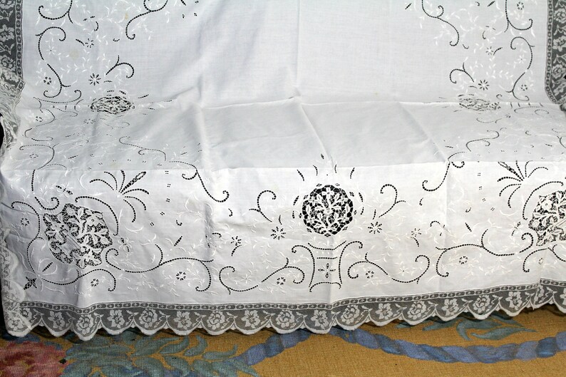 May include: White tablecloth with a delicate lace border and intricate embroidered floral designs in black thread.