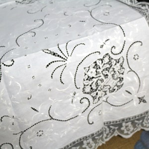 May include: White cotton tablecloth with delicate black embroidered floral designs. The tablecloth has a scalloped lace border.