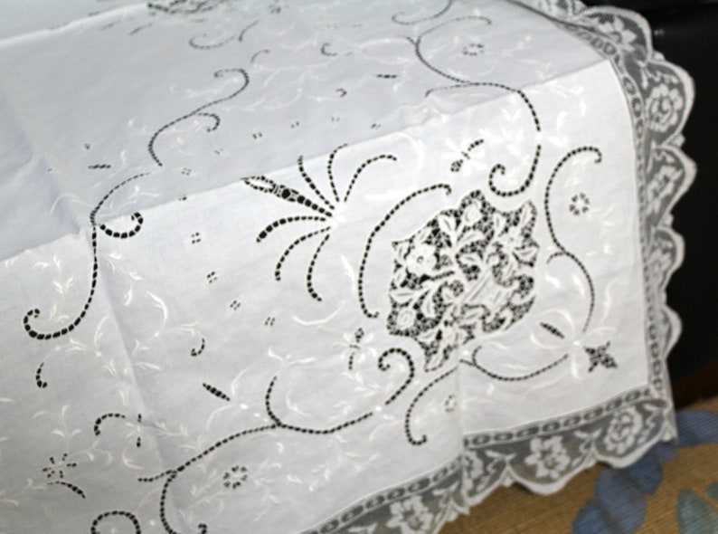 May include: White cotton tablecloth with delicate black embroidered floral designs. The tablecloth has a scalloped lace border.