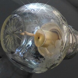 May include: A clear glass bottle with a silver top and a decorative etched design. The bottle has a small, round opening at the top and a clear glass tube extending from the top.