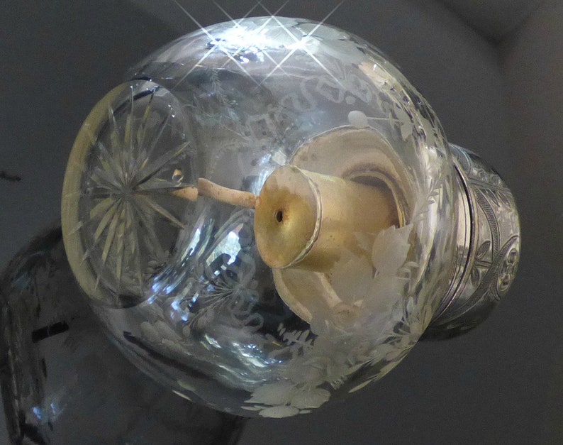 May include: A clear glass bottle with a silver top and a decorative etched design. The bottle has a small, round opening at the top and a clear glass tube extending from the top.