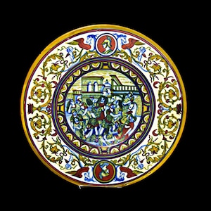 May include: A round ceramic plate with a colorful design. The center of the plate features a scene of people in a town, surrounded by a border of yellow, red, and blue floral patterns. The plate has a white background.