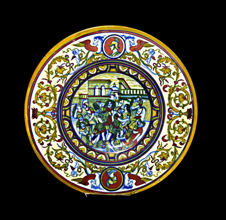 May include: A round ceramic plate with a colorful design. The center of the plate features a scene of people in a town, surrounded by a border of yellow, red, and blue floral patterns. The plate has a white background.