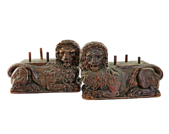 May include: A pair of antique wooden lion sculptures, carved in a dark brown colour. The lions are lying down with their heads facing forward. Each lion has a detailed mane and tail. The sculptures are in good condition with some signs of wear and tear.