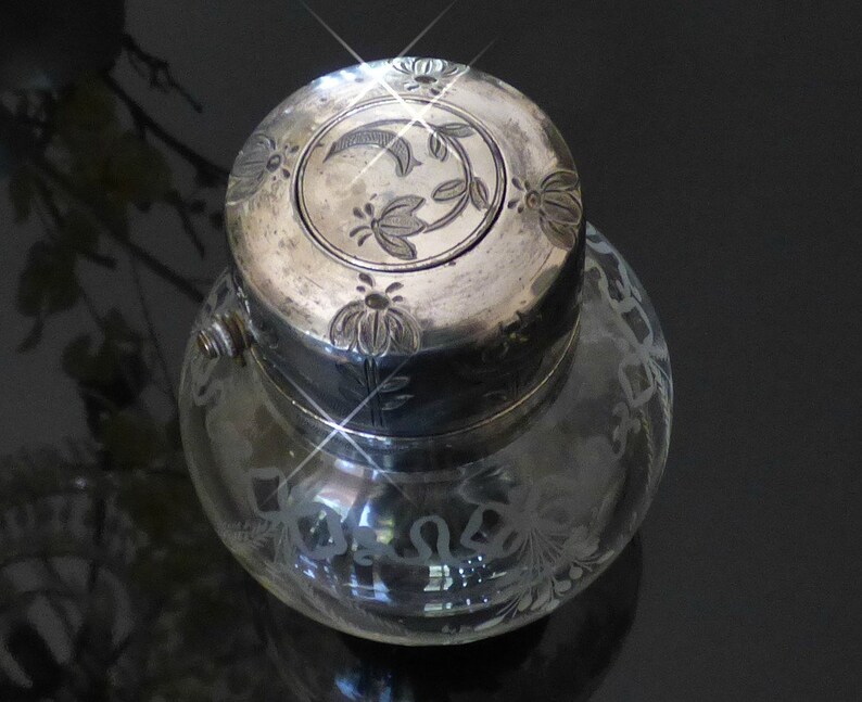 May include: A clear glass inkwell with a silver lid. The lid is decorated with an etched floral design. The inkwell is also decorated with an etched floral design.