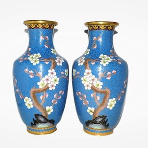 Puede incluir: Dos jarrones de cloisonné azul y dorado con un diseño floral. Los jarrones están decorados con un patrón floral blanco y rosa sobre un fondo azul. Los jarrones tienen un borde y una base dorados.