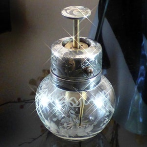 May include: A silver and glass bottle with a decorative top and etched floral design. The bottle has a silver cap with a small knob on the side.