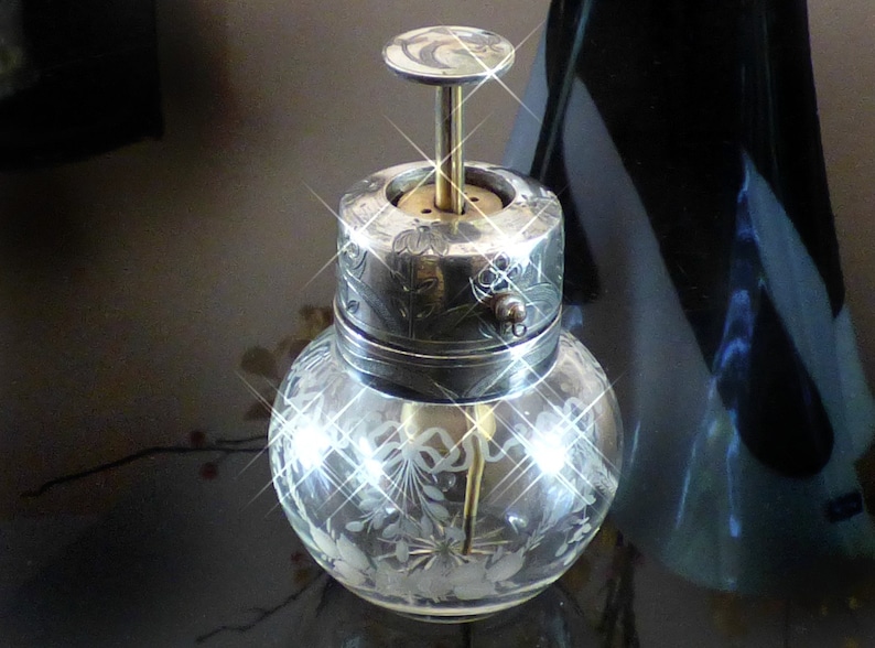 May include: A silver and glass bottle with a decorative top and etched floral design. The bottle has a silver cap with a small knob on the side.