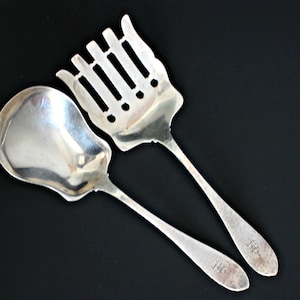 May include: A set of two silver serving utensils. The first utensil is a spoon with a rounded bowl and a long handle. The second utensil is a slotted spoon with a long handle. Both utensils have the same inscription on the handle: "T&B".