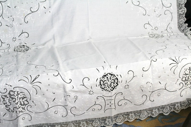 May include: White cotton tablecloth with delicate embroidered floral designs in black thread. The tablecloth has a scalloped edge with a lace trim.
