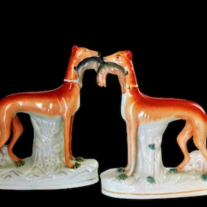 Puede incluir: Par de figuritas de cerámica de galgos marrones y blancos con un pez en la boca. Las figuritas están de pie sobre una base blanca con acentos verdes.