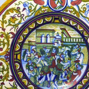 May include: A round ceramic plate with a colorful design. The center of the plate features a scene of people in a town square, with buildings in the background. The border of the plate is decorated with floral motifs and a blue and yellow pattern.