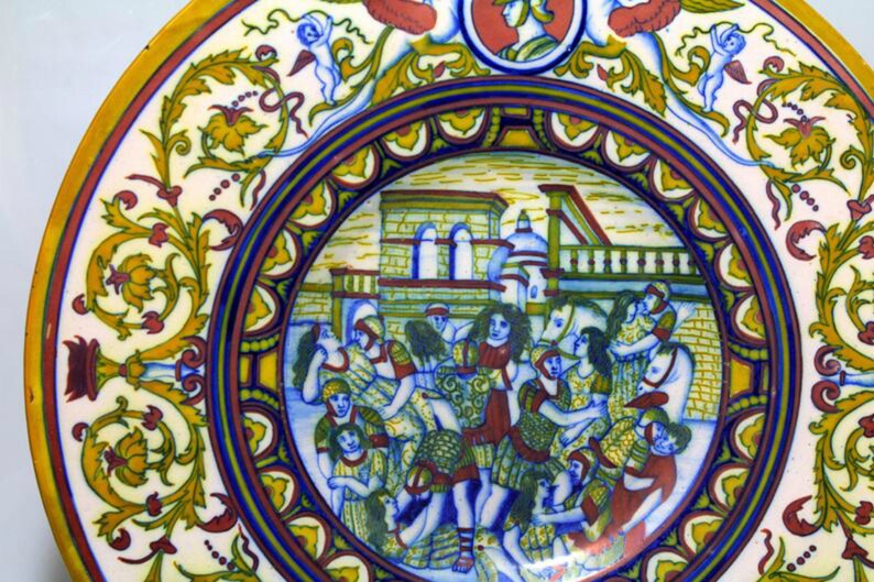 May include: A round ceramic plate with a colorful design. The center of the plate features a scene of people in a town square, with buildings in the background. The border of the plate is decorated with floral motifs and a blue and yellow pattern.