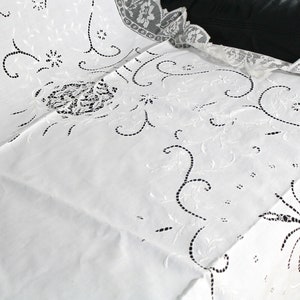 May include: White embroidered tablecloth with a delicate floral pattern. The tablecloth has a scalloped edge and is made of a fine, lightweight fabric.