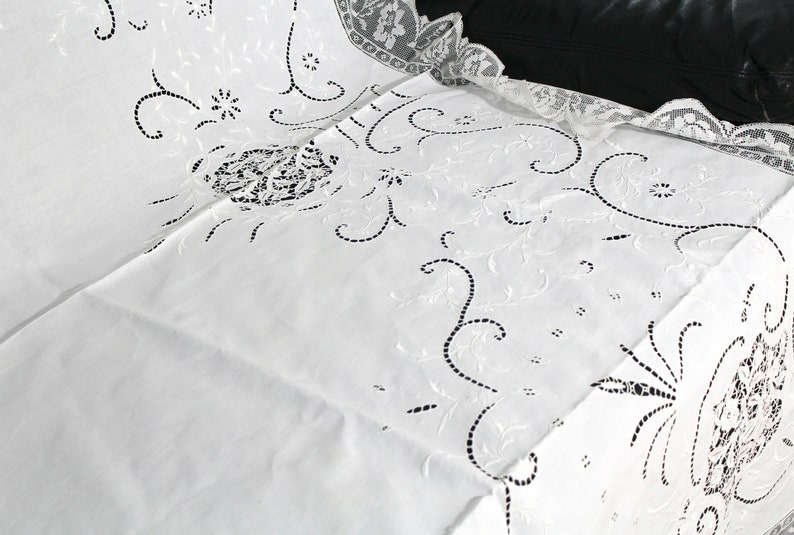 May include: White embroidered tablecloth with a delicate floral pattern. The tablecloth has a scalloped edge and is made of a fine, lightweight fabric.