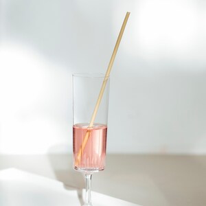 May include: A tall, clear glass filled halfway with a pink liquid, with a wooden straw resting inside. The glass sits on a white tray against a white background, with soft shadows.
