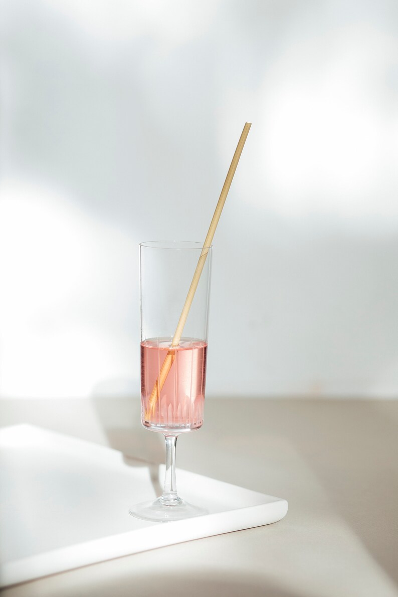 May include: A tall, clear glass filled halfway with a pink liquid, with a wooden straw resting inside. The glass sits on a white tray against a white background, with soft shadows.