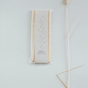 May include: A package of light-colored drinking straws with a beige paper label, featuring a minimalist leaf design. A dried wheat stalk is positioned to the right. The text on the label reads "STROH-TRINKHALME".