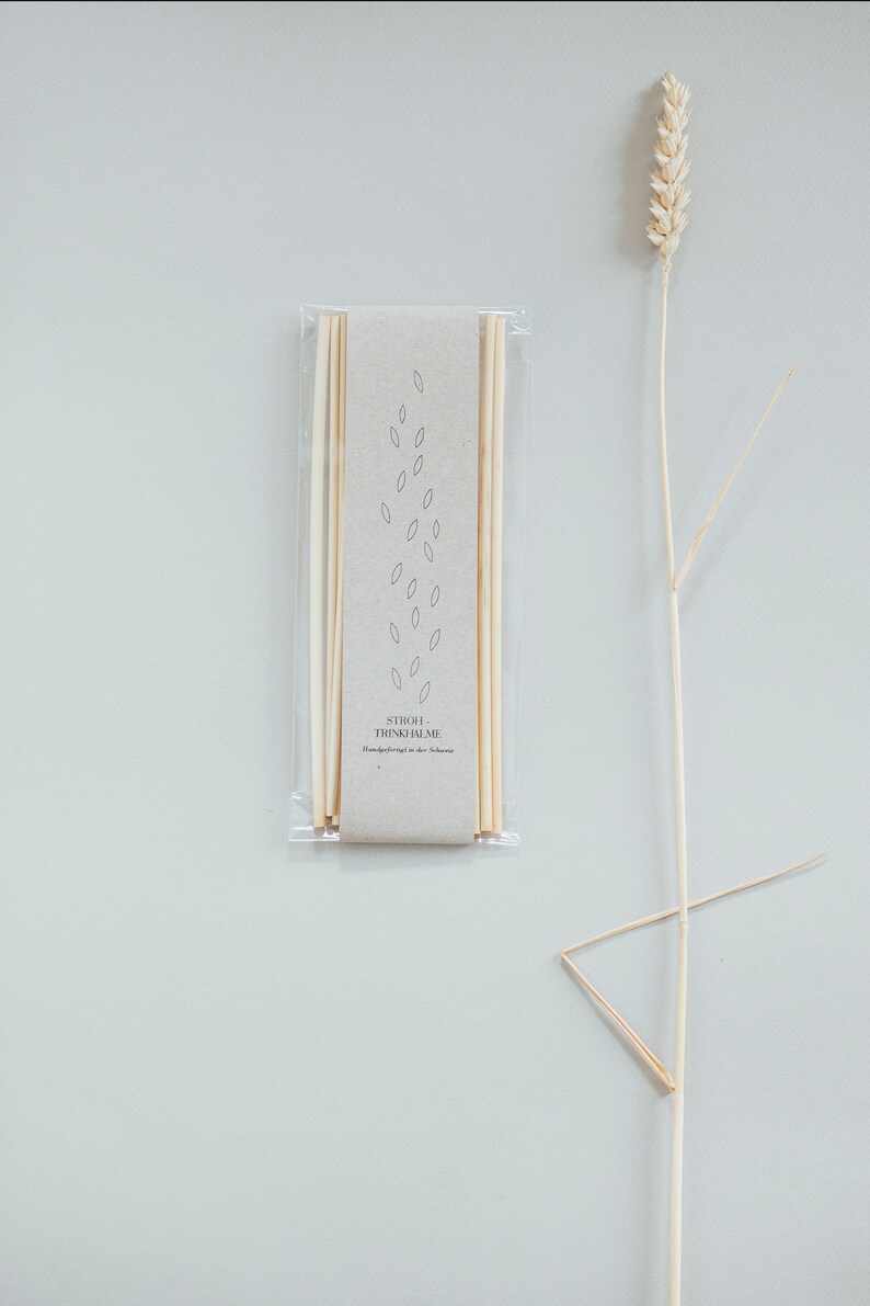 May include: A package of light-colored drinking straws with a beige paper label, featuring a minimalist leaf design. A dried wheat stalk is positioned to the right. The text on the label reads "STROH-TRINKHALME".