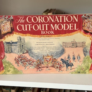 Puede incluir: Un libro modelo recortable vintage "The Coronation Cut-Out Model Book" con una cubierta roja. La cubierta presenta ilustraciones del Palacio de Buckingham, la Abadía de Westminster y una procesión de coronación.