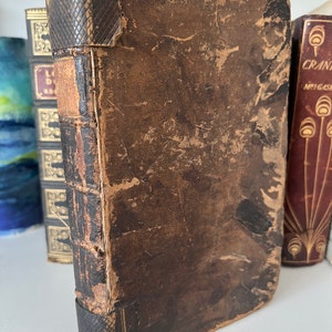 Op de afbeelding: Een vintage, bruin lederen gebonden boek met een versleten en verweerde uitstraling. Het boek heeft een decoratieve rug met een reliëf en staat licht schuin op een plank.
