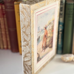 Può includere: Un libro d'epoca con un'illustrazione colorata sulla copertina, raffigurante una persona vicino al mare. Il libro ha un dorso e bordi dorati e color crema con motivi. Altri libri antichi sono visibili sullo sfondo.