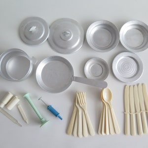 Puede incluir: Un juego de utensilios de cocina y utensilios de cocina de juguete hechos de aluminio. El juego incluye una olla, una sartén, una tapa, un plato, un tazón, una cuchara, un tenedor y un cuchillo. Los utensilios son todos de color plateado.