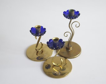 May include: Three brass candle holders with blue glass flower accents. The candle holders are in different sizes and have a decorative, swirling design.