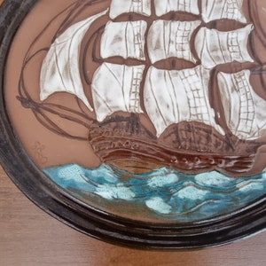 Puede incluir: Una placa de pared de cer&aacute;mica marr&oacute;n con una imagen en relieve de un velero blanco con velas marrones y un oc&eacute;ano azul. La placa tiene un marco marr&oacute;n oscuro.
