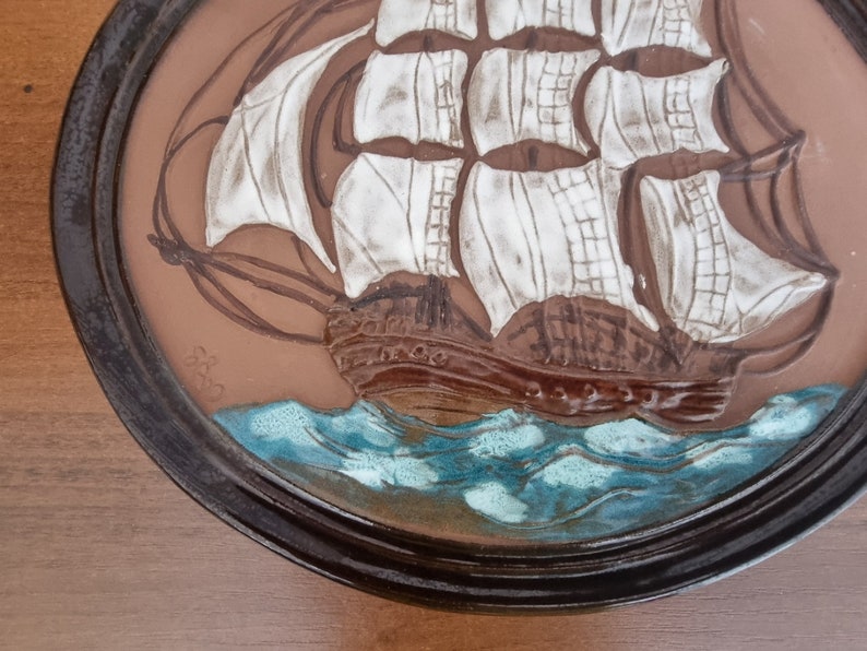 Puede incluir: Una placa de pared de cer&aacute;mica marr&oacute;n con una imagen en relieve de un velero blanco con velas marrones y un oc&eacute;ano azul. La placa tiene un marco marr&oacute;n oscuro.