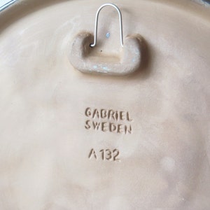 Puede incluir: Una placa de pared de cer&aacute;mica beige con un bucle de suspensi&oacute;n en la parte posterior. La placa tiene el texto "Gabriel Sweden A 132" estampado en la parte posterior.