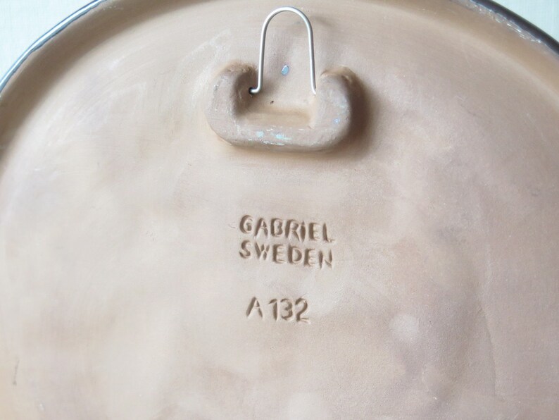 Puede incluir: Una placa de pared de cer&aacute;mica beige con un bucle de suspensi&oacute;n en la parte posterior. La placa tiene el texto "Gabriel Sweden A 132" estampado en la parte posterior.