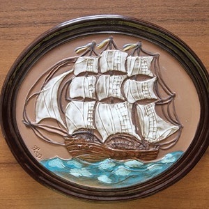Puede incluir: Una placa de pared de cer&aacute;mica marr&oacute;n con una imagen en relieve de un barco de vela blanco con velas marrones y un casco marr&oacute;n. El barco navega en aguas azules con olas blancas. La placa est&aacute; enmarcada en un marco de madera marr&oacute;n oscuro.