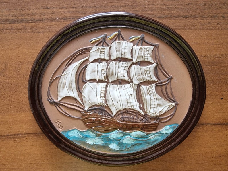 Puede incluir: Una placa de pared de cer&aacute;mica marr&oacute;n con una imagen en relieve de un barco de vela blanco con velas marrones y un casco marr&oacute;n. El barco navega en aguas azules con olas blancas. La placa est&aacute; enmarcada en un marco de madera marr&oacute;n oscuro.