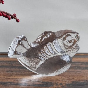 Pode incluir: Uma escultura de vidro transparente de um peixe com uma superfície texturizada detalhada. O peixe está posicionado com a cabeça inclinada para cima e a boca ligeiramente aberta.