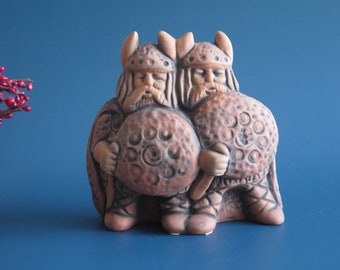 May include: Two ceramic Viking figures holding round shields with raised circular designs. The figures are brown and have beards and helmets with horns.
