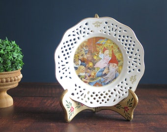 May include: White porcelain plate with a lacy edge, featuring a Christmas scene with a gnome and children. The plate is on a decorative stand.