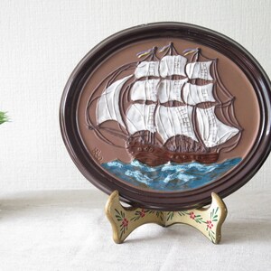 Puede incluir: Una placa de pared de cer&aacute;mica marr&oacute;n con una imagen en relieve de un barco de vela blanco con velas marrones y un oc&eacute;ano azul. La placa est&aacute; en un soporte decorativo con dise&ntilde;os florales.