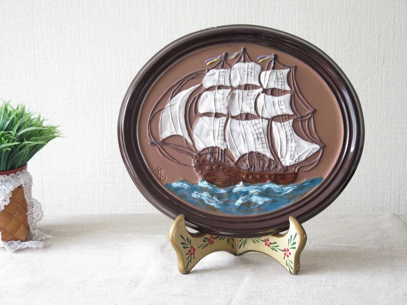 Puede incluir: Una placa de pared de cer&aacute;mica marr&oacute;n con una imagen en relieve de un barco de vela blanco con velas marrones y un oc&eacute;ano azul. La placa est&aacute; en un soporte decorativo con dise&ntilde;os florales.