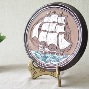 Puede incluir: Un plato de cer&aacute;mica marr&oacute;n con un dise&ntilde;o en relieve de un barco de vela blanco con m&aacute;stiles y velas marrones. El plato est&aacute; sobre un soporte dorado con flores pintadas de rojo y verde.