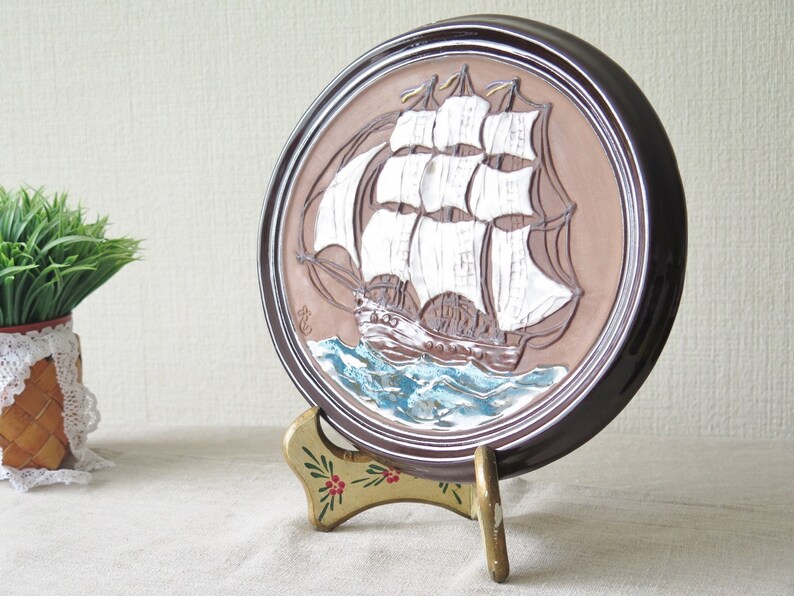Puede incluir: Un plato de cer&aacute;mica marr&oacute;n con un dise&ntilde;o en relieve de un barco de vela blanco con m&aacute;stiles y velas marrones. El plato est&aacute; sobre un soporte dorado con flores pintadas de rojo y verde.