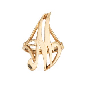 May include: A gold-colored ring featuring the letter "A" in a stylized, open design. The ring has a delicate, ornate appearance with curved lines and a polished finish. The ring is likely made of metal and is set against a white background.