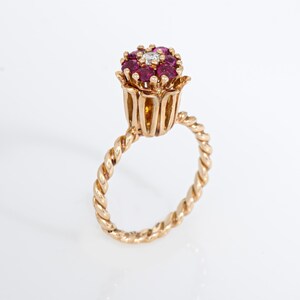 May include: A gold ring featuring a floral design. The flower-shaped top is adorned with small, round, red gemstones and a single, central diamond. The ring's band has a twisted rope design.
