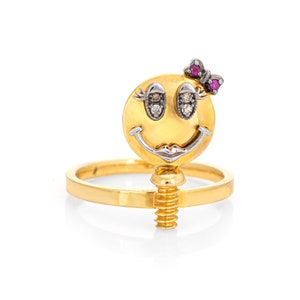May include: A gold ring with a smiley face charm. The smiley face has diamond eyes and a pink bow on top. The ring is attached to the charm with a screw.