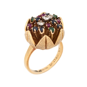 May include: A gold ring featuring a flower-shaped setting. The flower is crafted from gold and has a textured finish. The flower is adorned with an array of gemstones, including rubies, sapphires, emeralds, and diamonds. The ring is inscribed with the words "Van Cleef & Arpels".