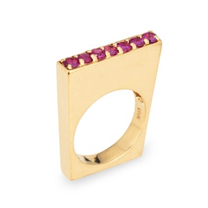 Può includere: Un anello d'oro con una fascia quadrata e una fila di pietre preziose rosa incastonate sul bordo superiore. L'anello è contrassegnato "18K" all'interno.