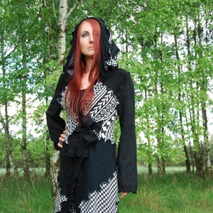 May include: A woman wearing a black and white patterned hooded cardigan with a ruffled hem. The cardigan is made of a soft, knitted fabric and has a relaxed fit. The woman is standing in a woodland setting.