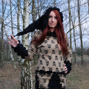 May include: A woman with long red hair is wearing a black hooded sweater with a skull and crossbones pattern. The sweater has a ruffled trim and the hood is pulled up over her head. She is standing in a forest with birch trees in the background.