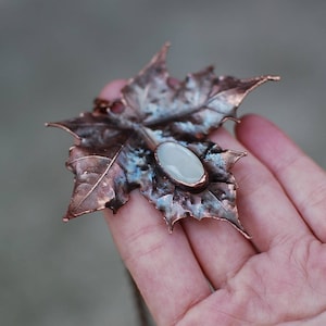 Puede incluir: Un colgante de hoja de cobre con una piedra ovalada blanca en el centro. La hoja es texturizada y tiene un aspecto rústico.