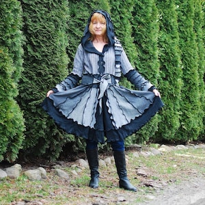 Könnte beinhalten: Eine Frau trägt einen grauen und schwarzen Kapuzenmantel mit einem Rüschen-Saum. Der Mantel ist aus verschiedenen Stoffen gefertigt und hat ein einzigartiges Design. Sie steht vor einer Reihe von immergrünen Bäumen.